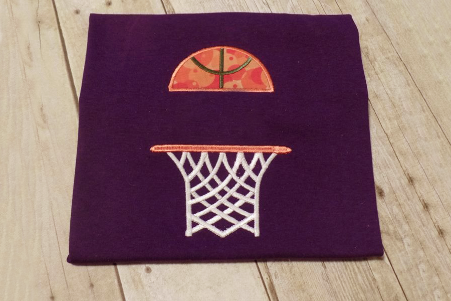 A purple square of fabric sits on a wood background. Embroidered onto the fabric is a basketball hoop with half an applique basketball above, with a split space in between the two.