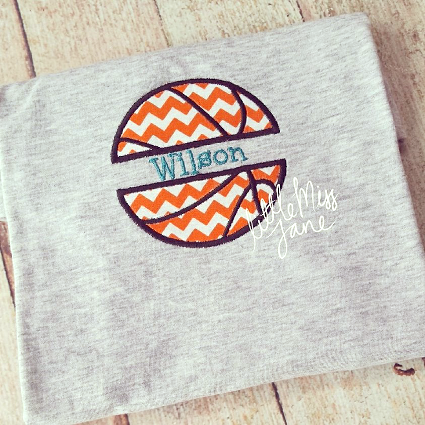 A heather grey folded shirts sits on a wooden backdrop. Embroidered onto the shirt is a basketball applique with a split in the middle, done in orange chevron applique fabric. The customer has added the embroidered word "Wilson" into the split space. (Text is not included). Little Miss Jane watermark is on the photo.