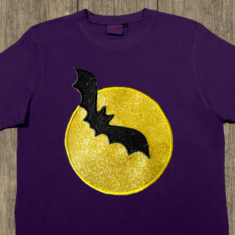 A purple child's shirt sits on a wood backdrop. Embroidered on the shirt are a glittery black applique bat flying against a glittery yellow applique moon.