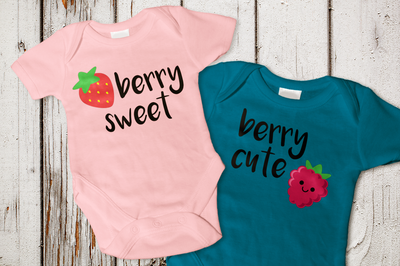 Two baby onesies on a weathered wood background. Left onesie has a strawberry and says "berry sweet." Right onesie has a smiling raspberry and says "berry cute."