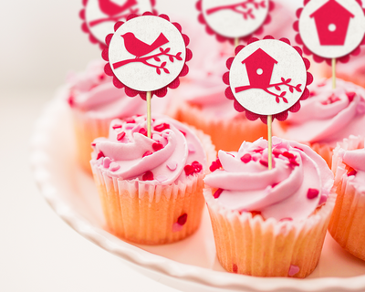 Cupcakes with pink frosting. Each has a cupcake topper that is a circle with a fuchsia colored scallop border and image in the center. The center images are a birdhouse on a branch, a branch, a bird on a branch, and a birdhouse.