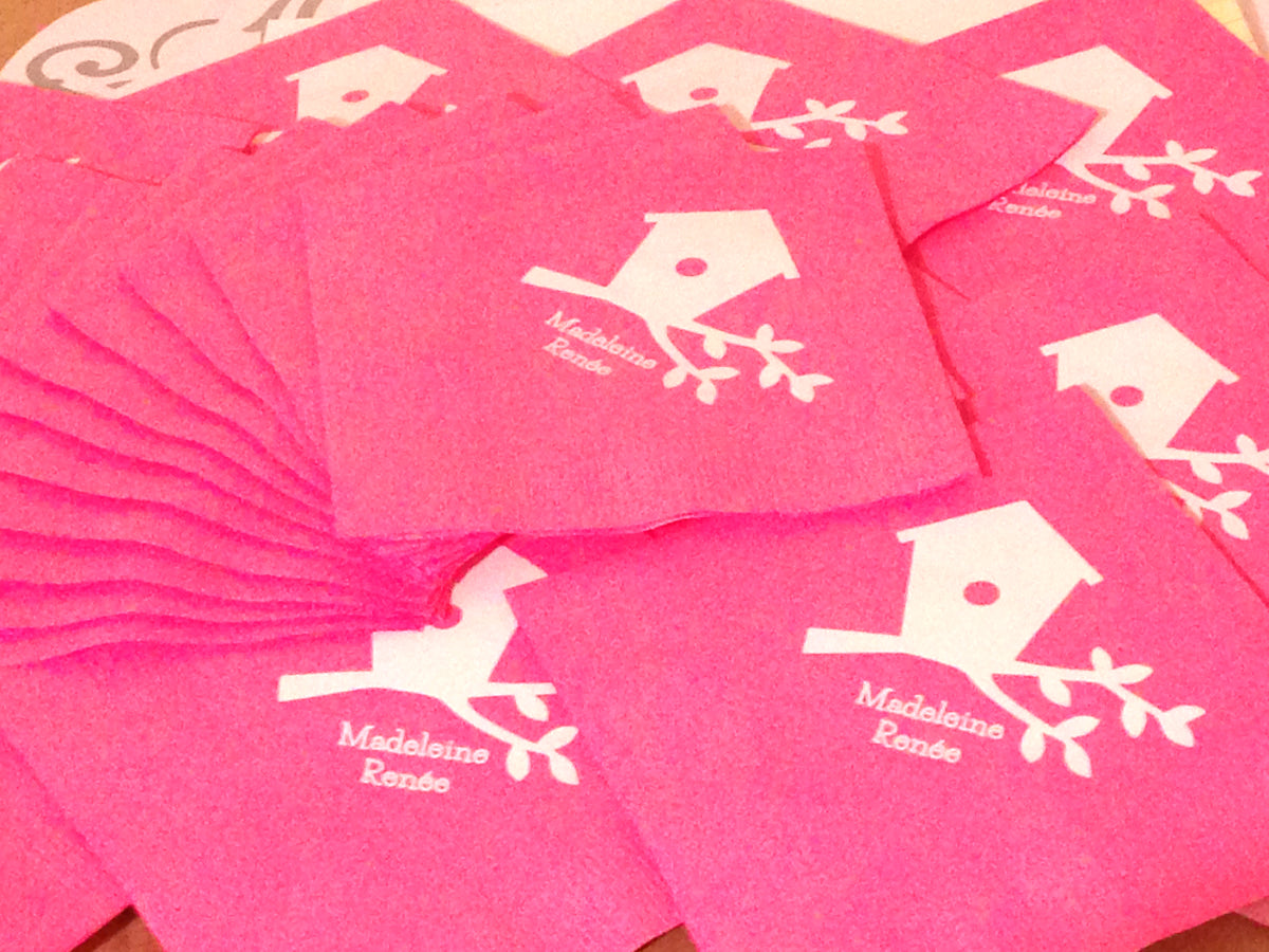 Pink napkins are fanned out. Each has a white image of a birdhouse on a branch with a name added below.