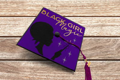 A graduation cap on a wood background. The design on the cap has a cameo style profile of a black woman from the neck up with a scarf and an afro and the words "Black Girl Magic." She also holds a wand and there are stars and a swirl around the wand.