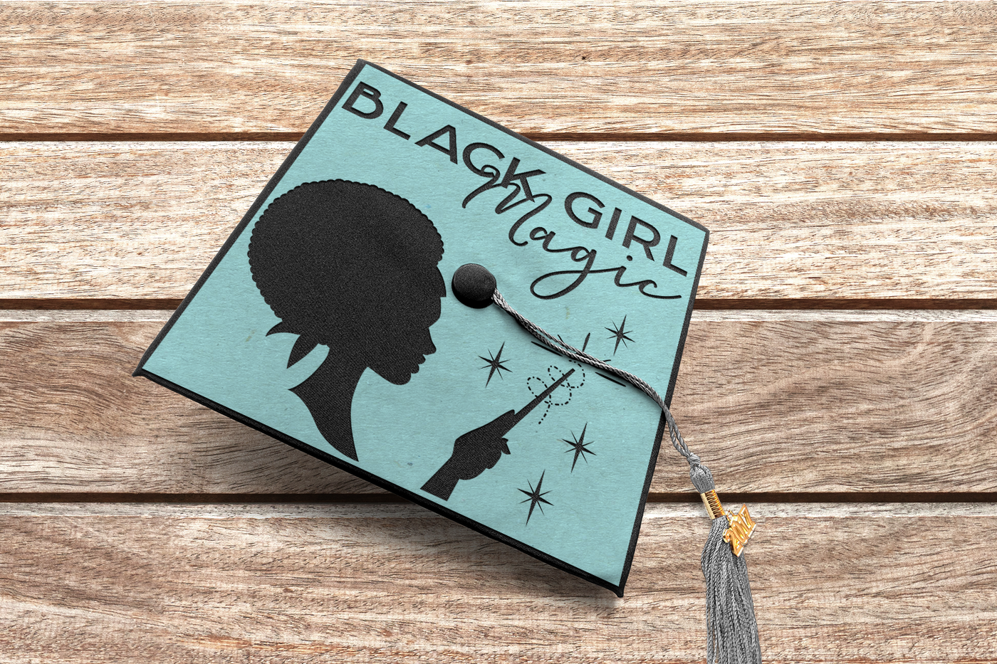 A graduation cap on a wood background. The design on the cap has a cameo style profile of a black woman from the neck up with a scarf and an afro and the words "Black Girl Magic." She also holds a wand and there are stars and a swirl around the wand.