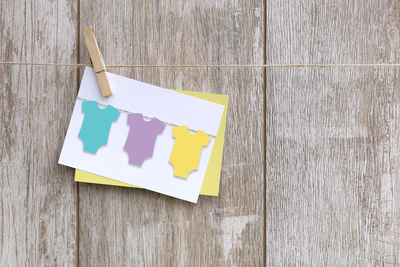 White card with 3 pastel bodysuits on a string. Behind the card is a yellow envelope. Card is pinned to a clothesline against a wood background.