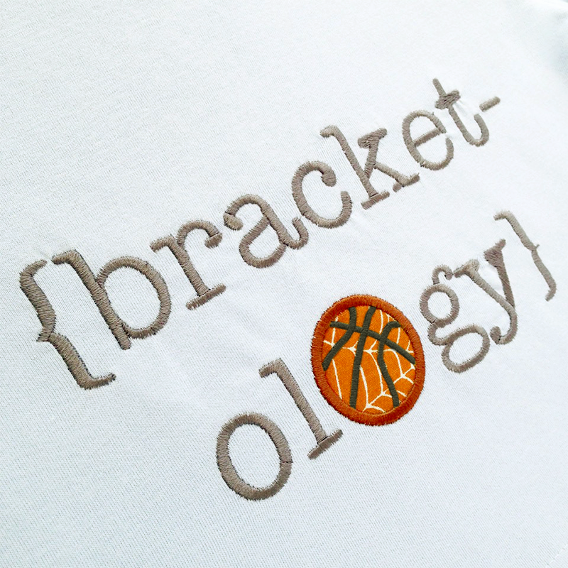 A pale blue fabric is embroidered in silver with the word "{bracket-ology}." One of the Os is an applique basketball.