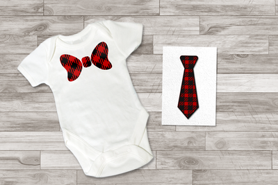 A white baby onesie and card lay on a wood background. The onesie has a buffalo plaid bow tie, the card has a layered paper buffalo plaid necktie. Both ties are in red and black.