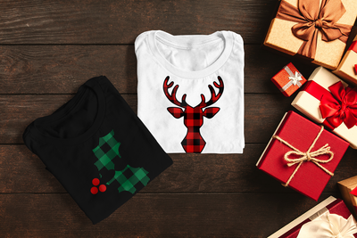 2 folded tees on a wood surface with presents to the side. One tee has a buffalo plaid sprig of holly, the other has a buffalo plaid reindeer bust.