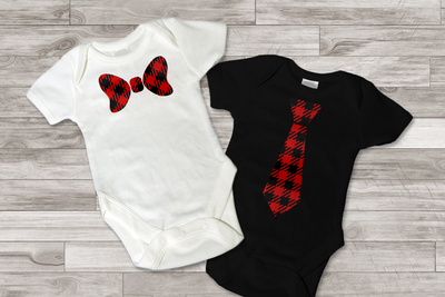 Two baby onesies on a wood background. One has a neck tie, the other has a bow tie. Both are red and black with a buffalo plaid pattern.