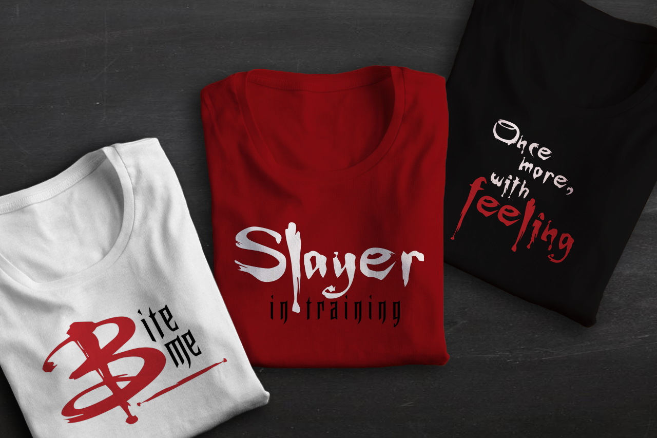Three folded shirts in white, red, and black. Each shirt has a design in those same colors. The left says "Bite me," the middle says "Slayer in training," and the right says "Once more, wit feeling."