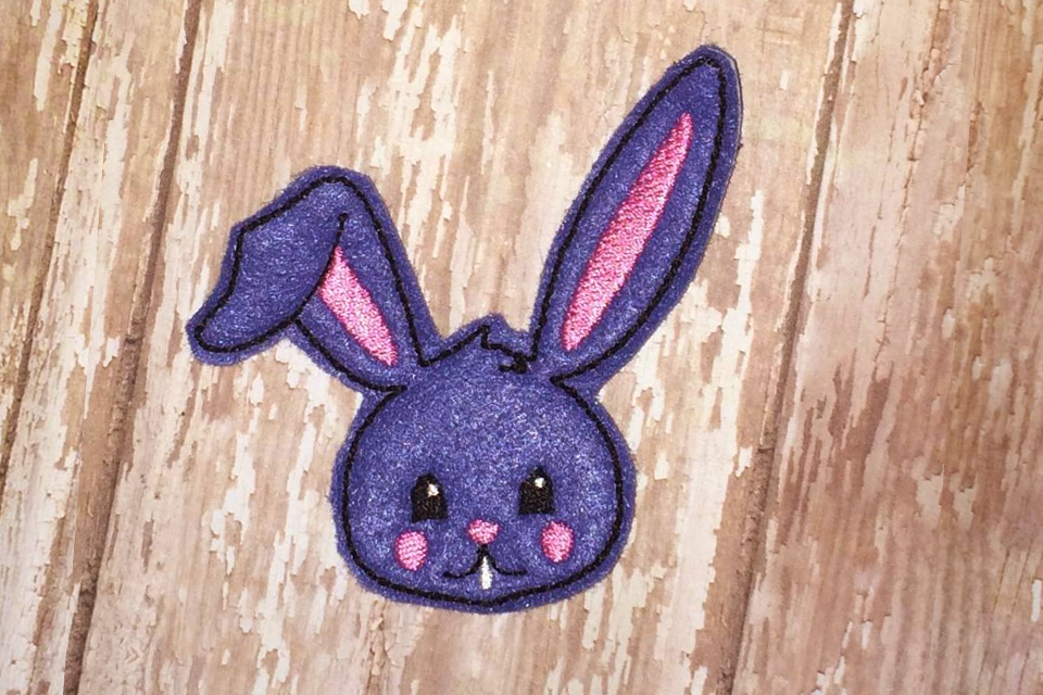 Purple Easter bunny face feltie with one ear partially flopped down.
