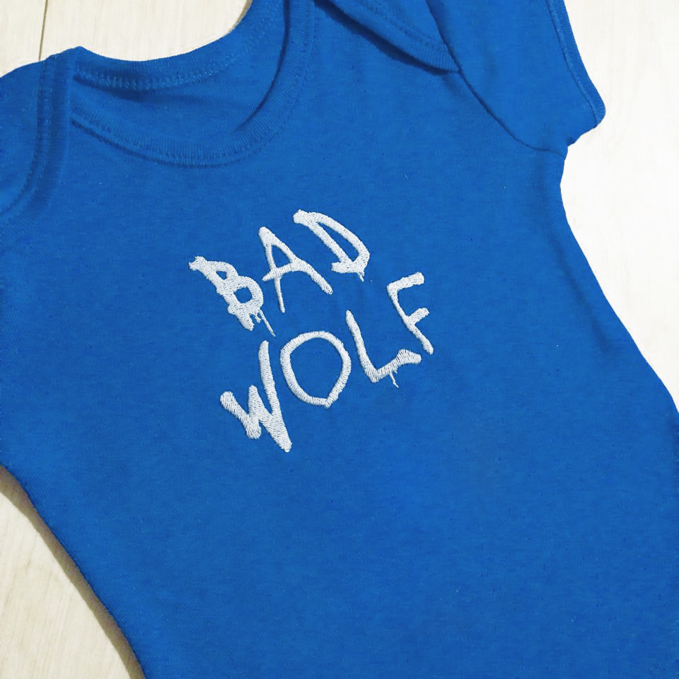 A royal blue baby onesie lays on a white surface. Embroidered on it in white are graffiti style letters that say "bad wolf."