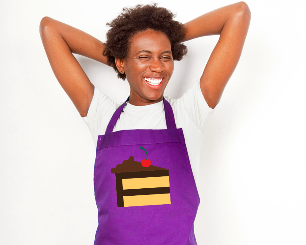 A laughing black woman wears a white tee and purple apron. On the apron is a yellow cake with chocolate frosting and a cherry design.