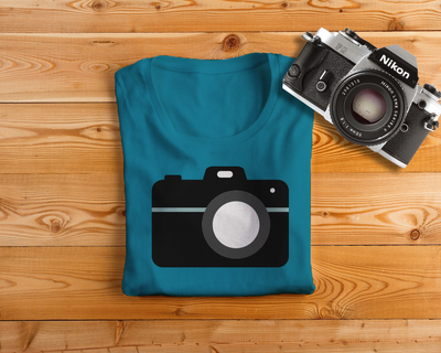 A teal tee on a wood background with a Nikon camera to the side. The shirt has a DSLR camera design on it.