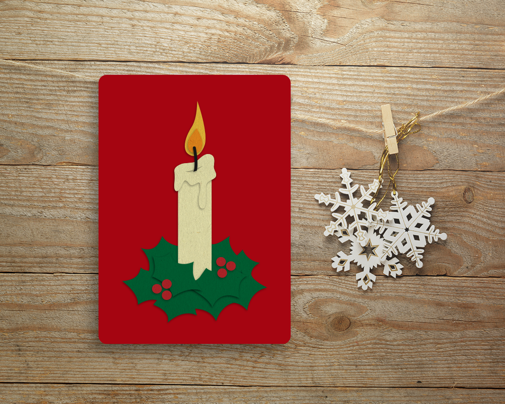 A red card on a wood background. Next to it are white and gold snowflake ornaments. Ont he card in layered paper is lit candle with dripping wax and holly around the base.