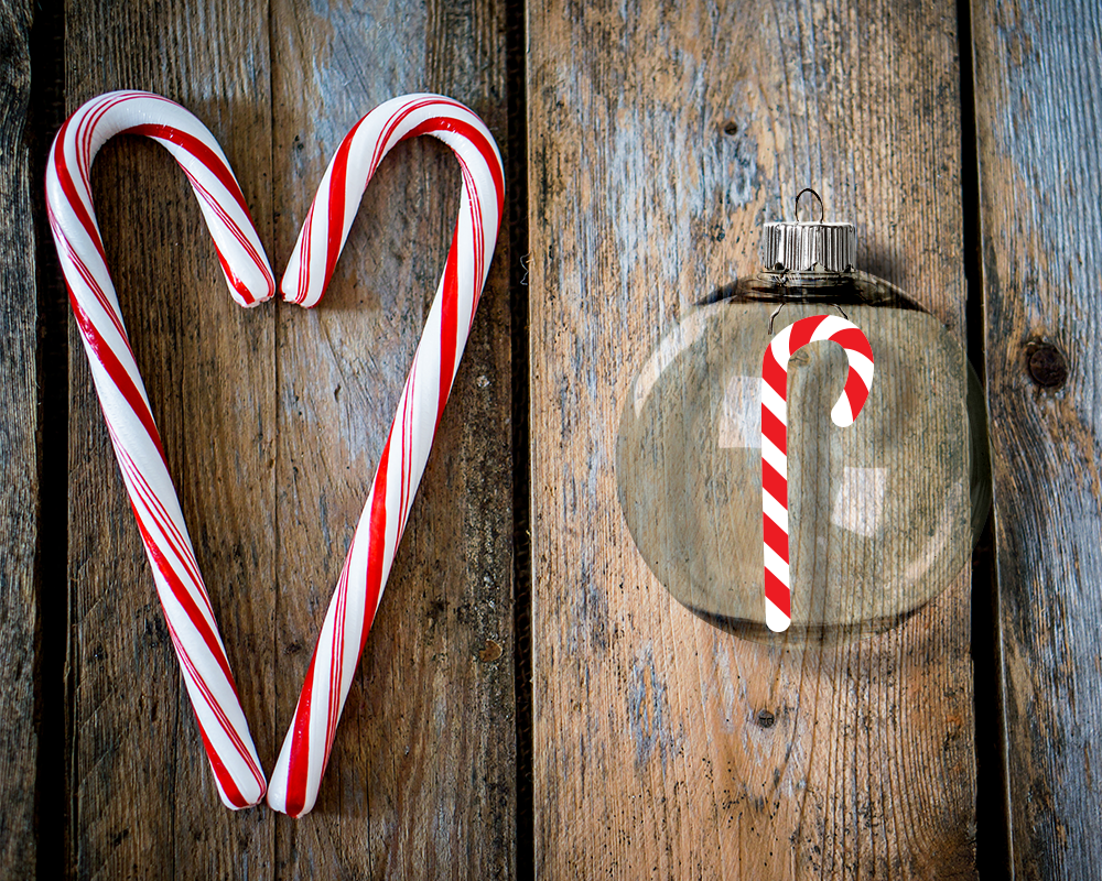 A clear round glass ornament sits on a wood background. Next to the ornament are 2 candy canes. The ornament has the design of a single candy cane.