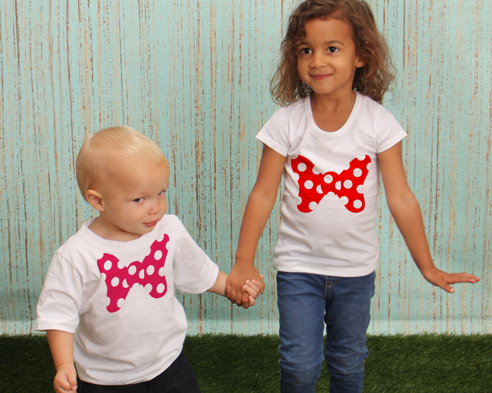 A black little girl and white-presenting toddler of color stand holding hands. Each has a white tee with an oversized bow with polkadots. The toddler's bow is hot pink and white, the child's bow is red and white.