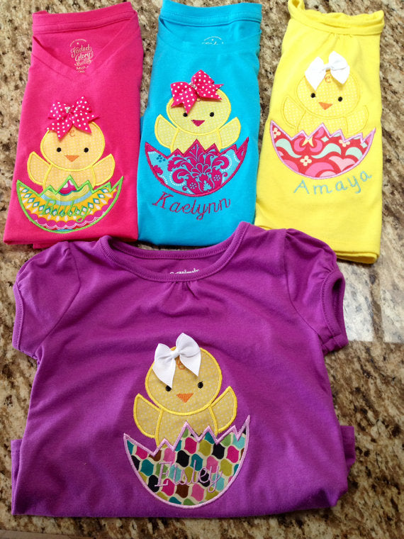 Five folded shirts. Each has the applique design of a chick popping out of a cracked egg.