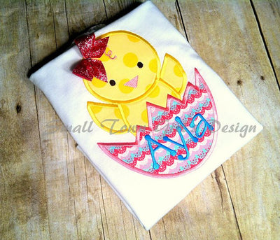 A folded tee with an applique design of a chick popping out of a cracked egg.