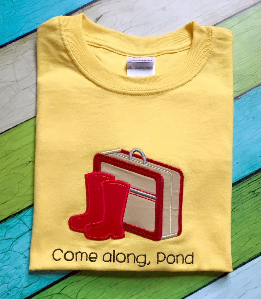 Yellow shirt with an applique suitcase and rain boots. Embroidered on the shirt it says, "Come along, Pond."