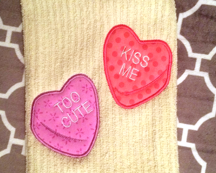 A pale yellow tea towel has the applique design of two candy conversation hearts. One says "too cute" and the other says "kiss me."