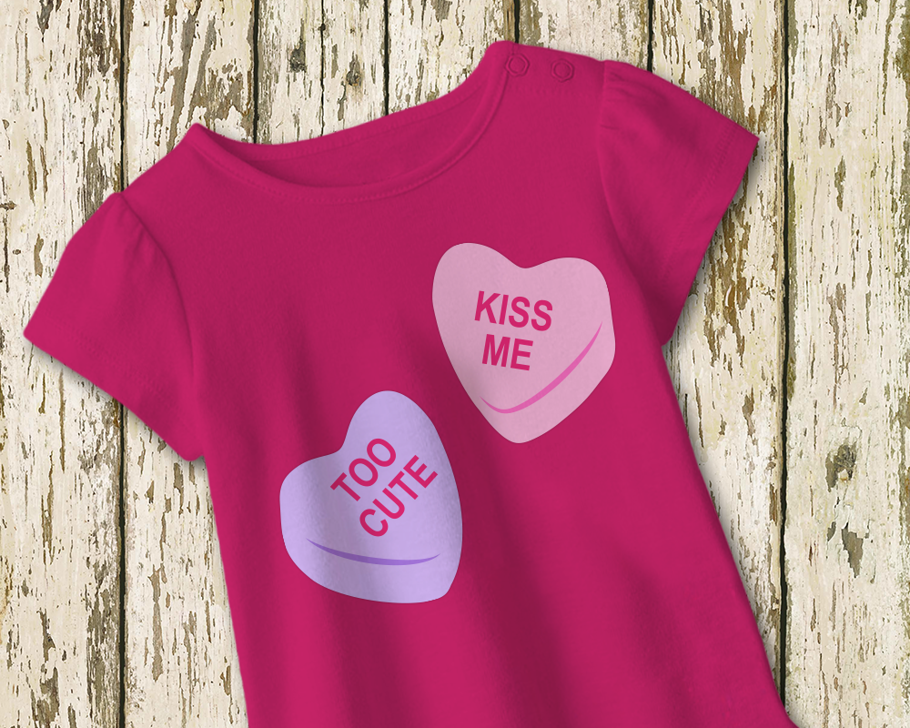 A dark pink tee with a design of 2 candy conversation hearts. One says "Too cute" and the other says "Kiss me."