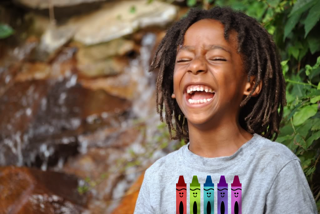 A black little boy is standing outside in front of a waterfall and laughing. He wears a heather grey tee with a design of 5 smiling crayons in different colors.
