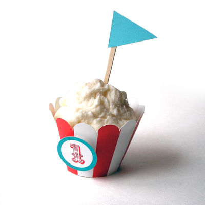 A vanilla frosted cupcake with a red and white striped scallop wrapper. A circle pops out from the cupcake that has a turquoise outline and red number 1 in the middle in circus style lettering. The cupcake has a turquoise triangle flag topper.
