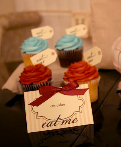 A table tent that is labeled "cupcakes" and says "eat me" at the bottom. The card is ivory with tan stripes and a burgundy bow. Behind it are cupcakes with tag toppers that say "eat me."