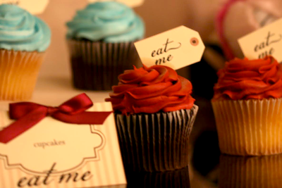 Frosted cupcakes that have tag toppers saying "eat me. " In front is an ivory table tent with tan stripes and a burgundy bow that is labeled "cupcakes" with "eat me" below.