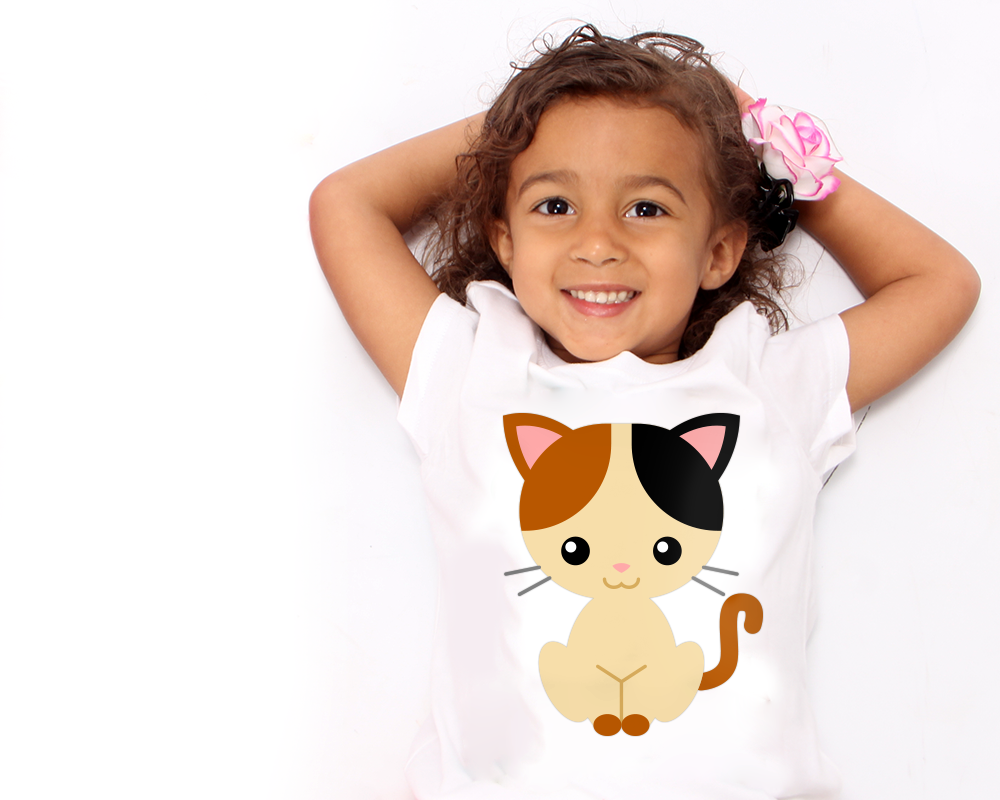 A little black girl lays on a white surface with her arms under her head. She is smiling and has a cute calico kitten on her white tee and a pink and white flower in her hair.