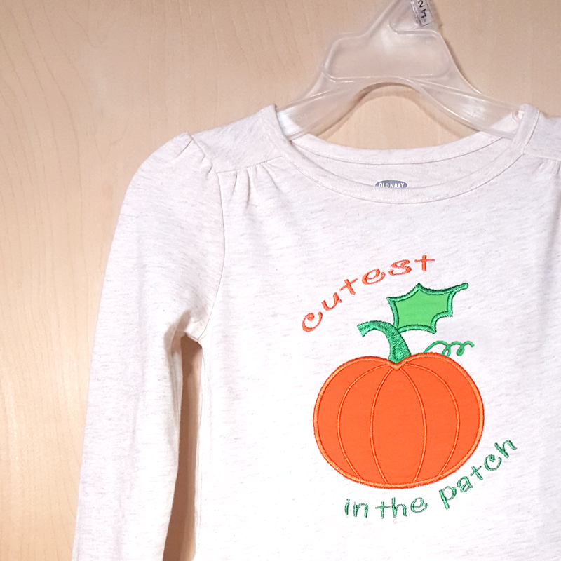 A white dress with an applique pumpkin. Around the pumpkin it says "cutest pumpkin in the patch."