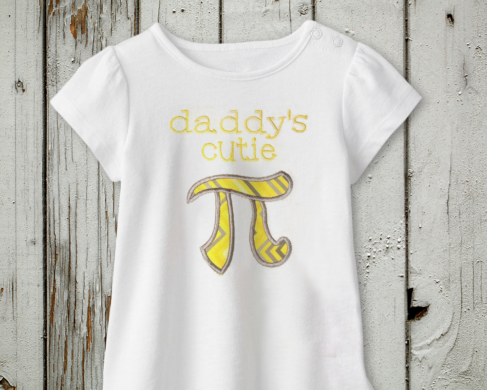 A white child's shirt. Embroidered onto the shirt it says "daddy's cutie" with a pi symbol applique below.