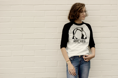 A white-presenting Latinx teen girl wears a black and white raglan tee. She stands in front of a white brick wall. On her shirt it says "Arches National Park" with an image of the arch rock formation.