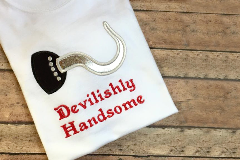 White folded shirt with applique design of a pirate's hook. Embroidered below it says "Devilishly Handsome."