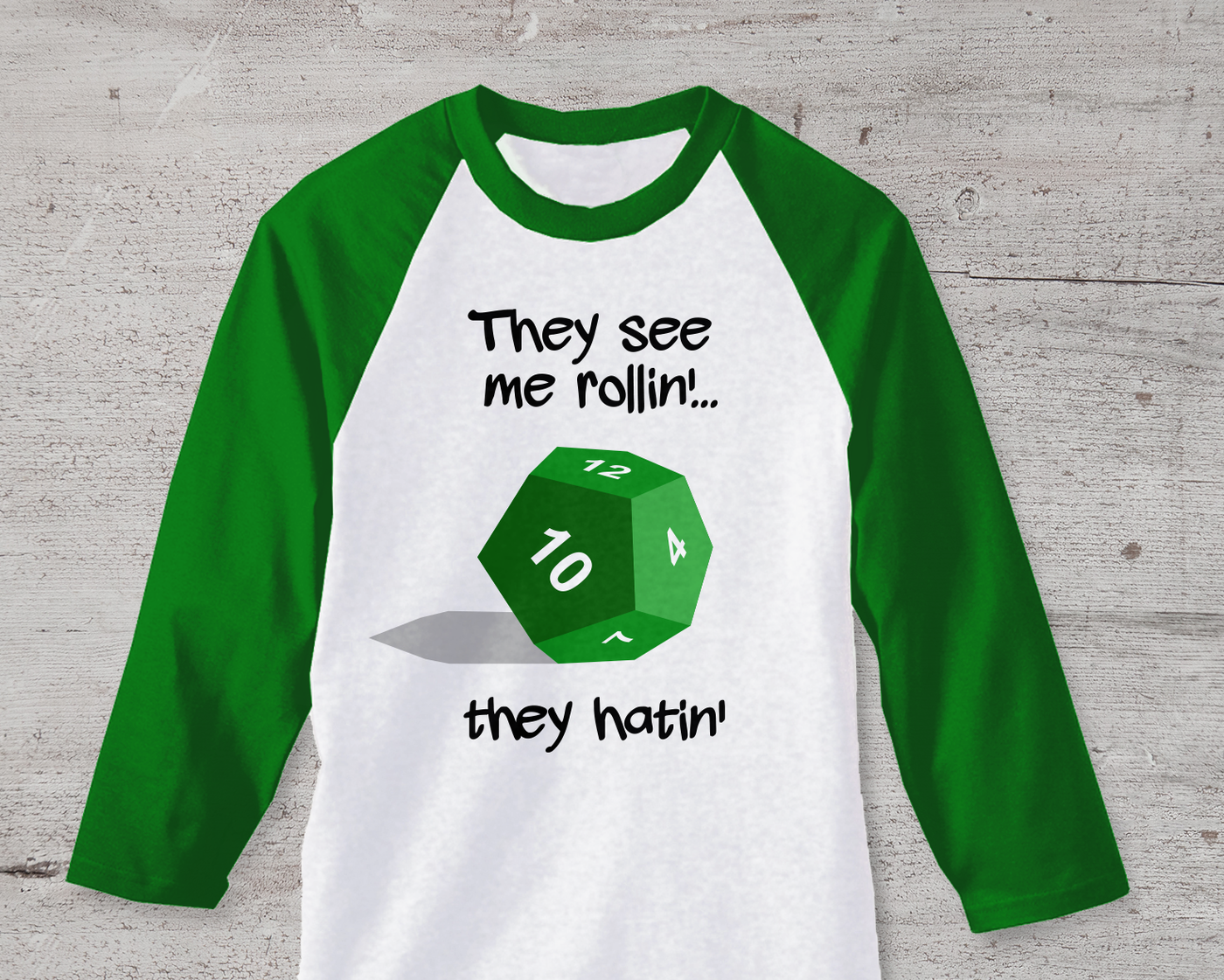 A green and white raglan tee sits on a cement background. There is a green 12-sided die in the center with the words "They see me rollin'... they hatin'" around it.