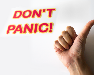 Letters with rounded edges in red with a yellow border made out of paper. Says "Don't Panic!" and a hand is making the hitchhiking gesture with a raised thumb.