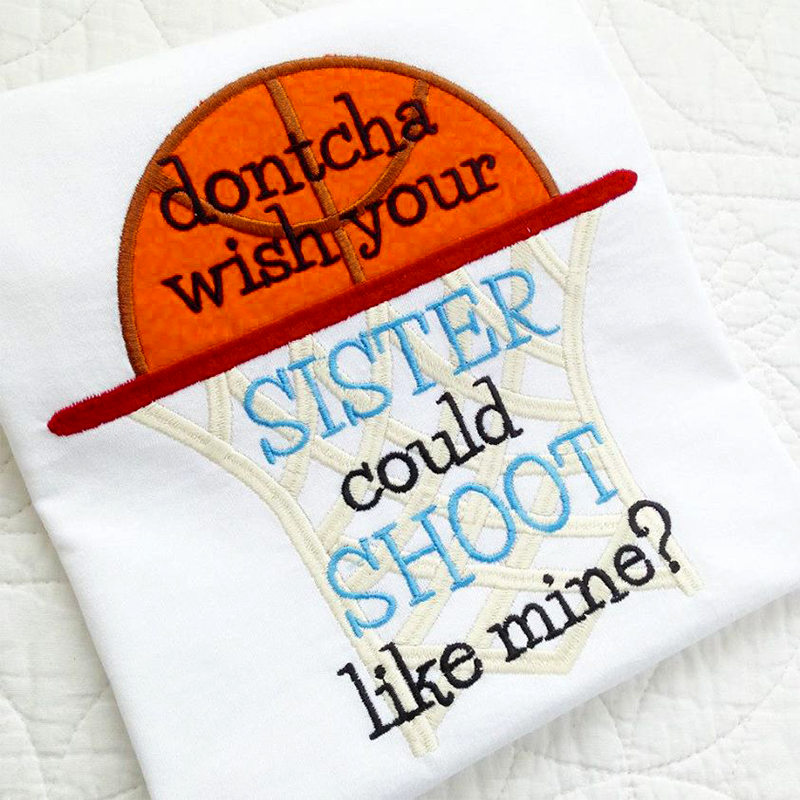 A white folded shirt with an applique basketball going through a hoop. Embroidered on top are the words "dontcha wish your SISTER could SHOOT like mine?"