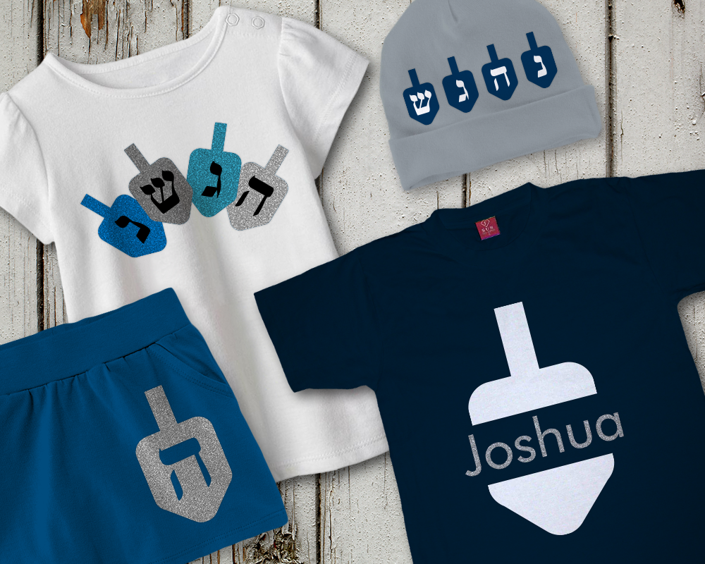 Children's clothing laid out. The skirt has a single dreidel. The hat has 4 dreidels, each with a different Hebrew letter. One shirt has a dreidel with a split in the middle and the name Joshua added (name/font not included). The other shirt has 4 dreidels fanned out, each with a different Hebrew letter.
