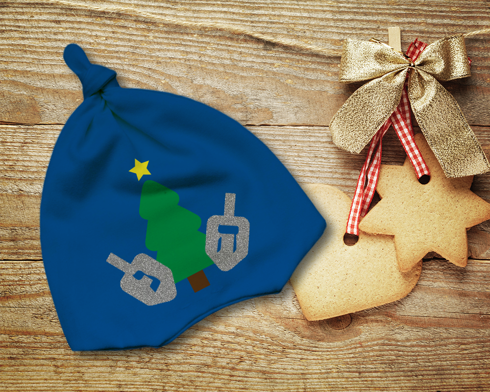 A blue knotted baby cap on a wood surface. Next to it are holiday ornaments that look like sugar cookies. The cap is decorated with a Christmas tree with 2 dreidels on either side.