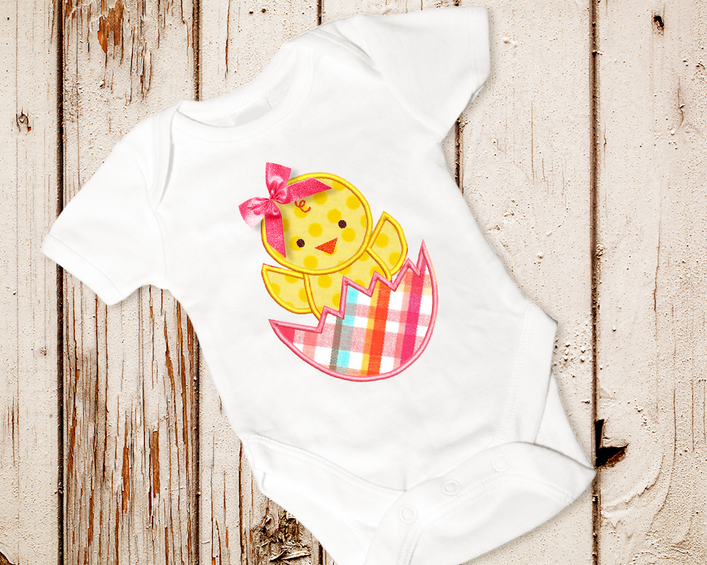 A baby onesie with a chick popping out of a cracked egg applique design. A real bow has been added to the chick's head.