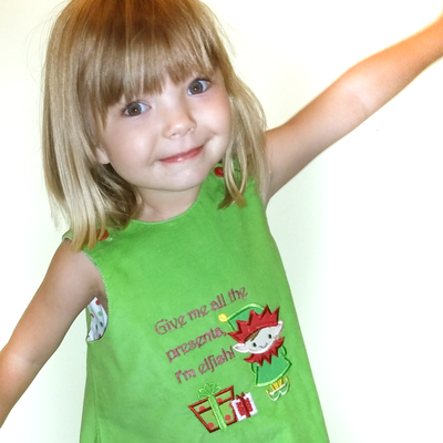 White little girl in a green dress. On the dress is an Elf applique with the words "Give me all the presents, I'm elfish!"