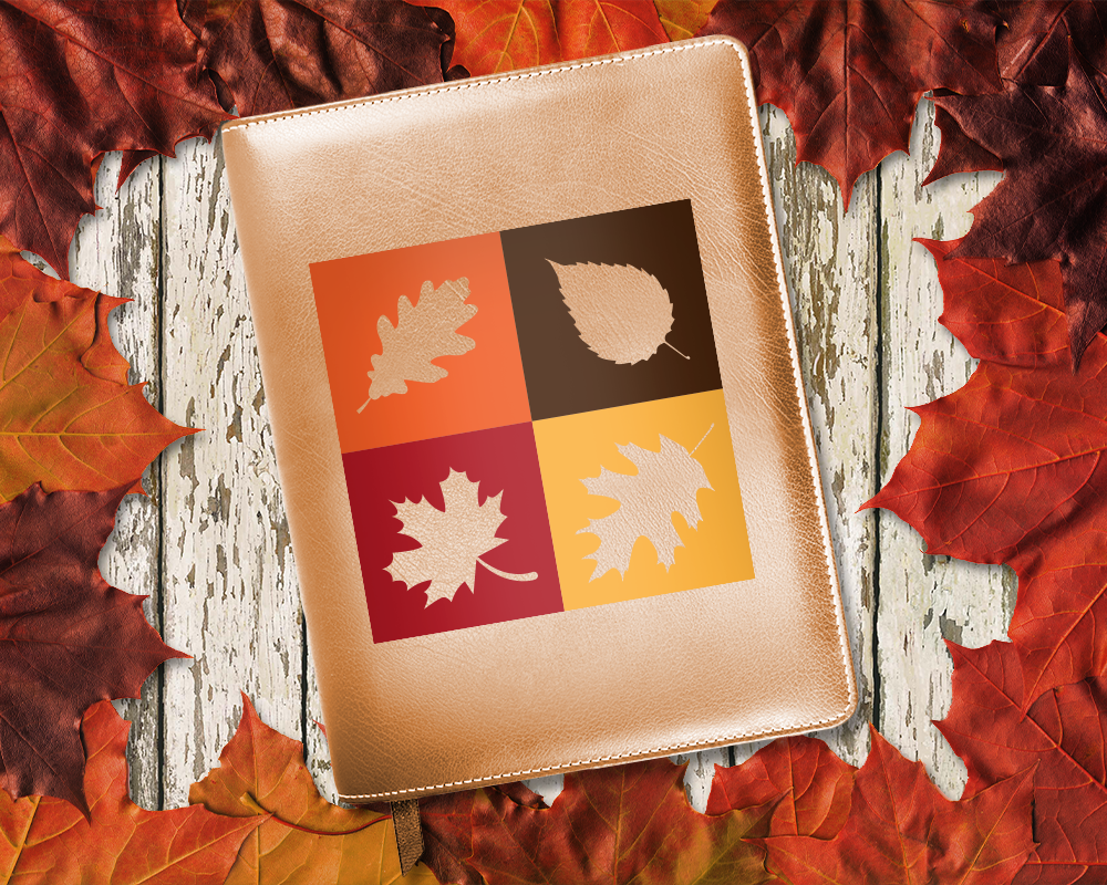 A copper colored planner surrounded by fall leaves. The design on the planner is 4 squares with leaf shapes as knockouts. The squares are in red, orange, yellow, and brown.