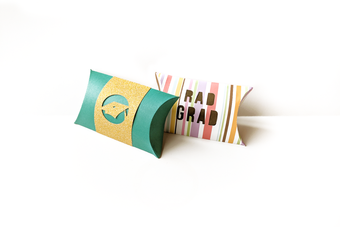 Two pillow boxes. One is turquoise with a gold band that has a grad cap cutout. The other is striped with cutout text reading "Rad grad."