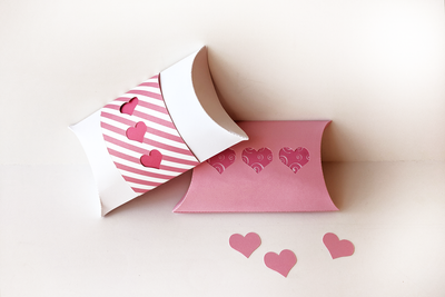 Two pillow boxes on a white background. The white box has a pink and white striped band with 3 heart cutouts revealing pink hearts. The other is pink with 3 heart cutouts revealing a darker pink patterned paper.