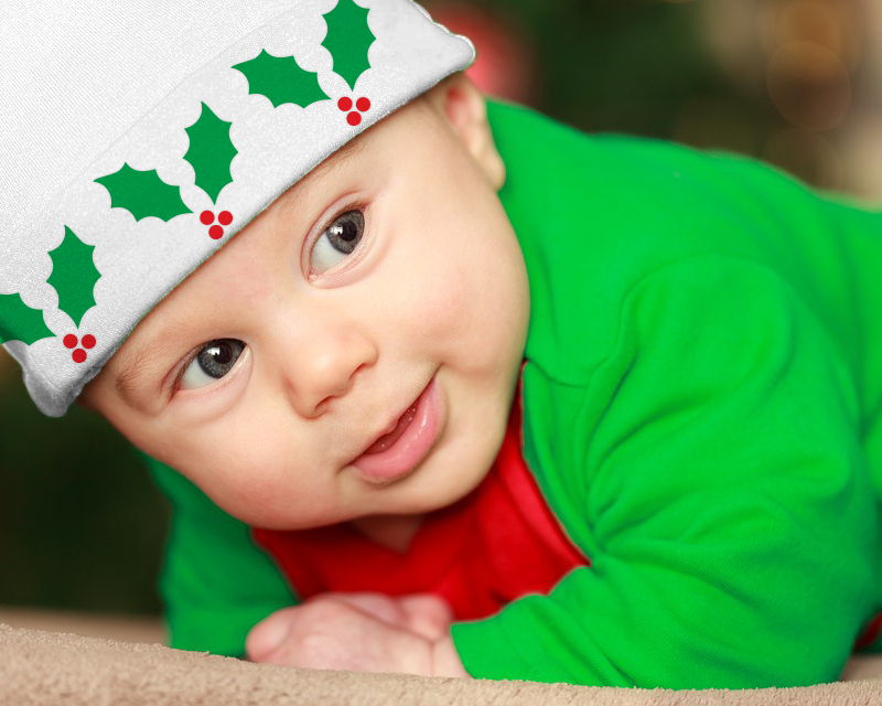 White baby wears a red and green outfit and white knit cap. The knit cap has 3 sprigs of holly on it.