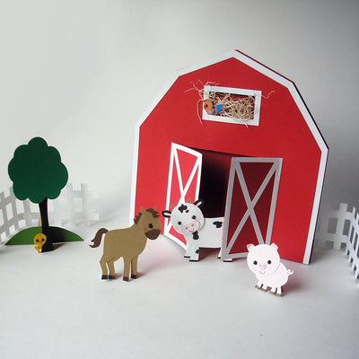 A paper barn with doors that swing out is standing up on a white background. Around the barn is a paper fence, tree, and various paper farm animals. (Animals, tree, and fence not included).