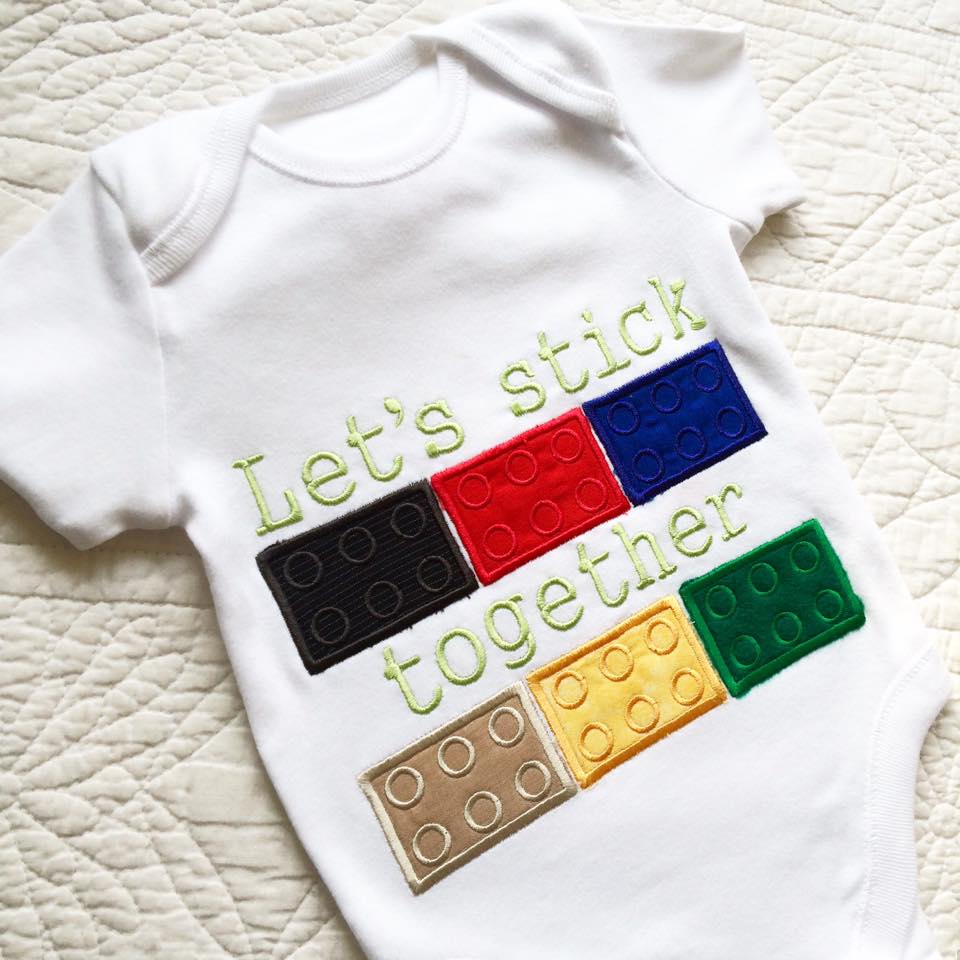 A white baby onesie on a white background. It has an applique design of building bricks on it with a row of bricks at the top and bottom with a split space in between. Above and between the bricks, the customer has embroidered the words "Let's stick together." (Words are not included as part of the design file.)