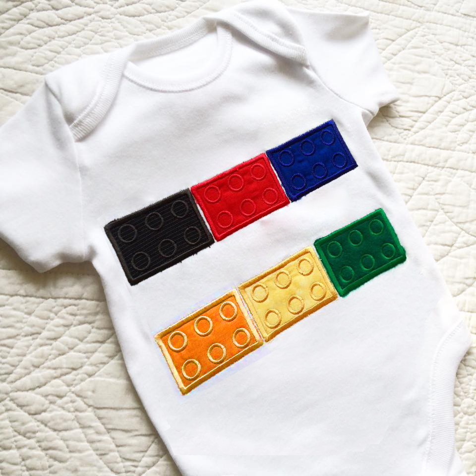 A white baby onesie on a white background. It has an applique design of building bricks on it with a row of bricks at the top and bottom with a split space in between.