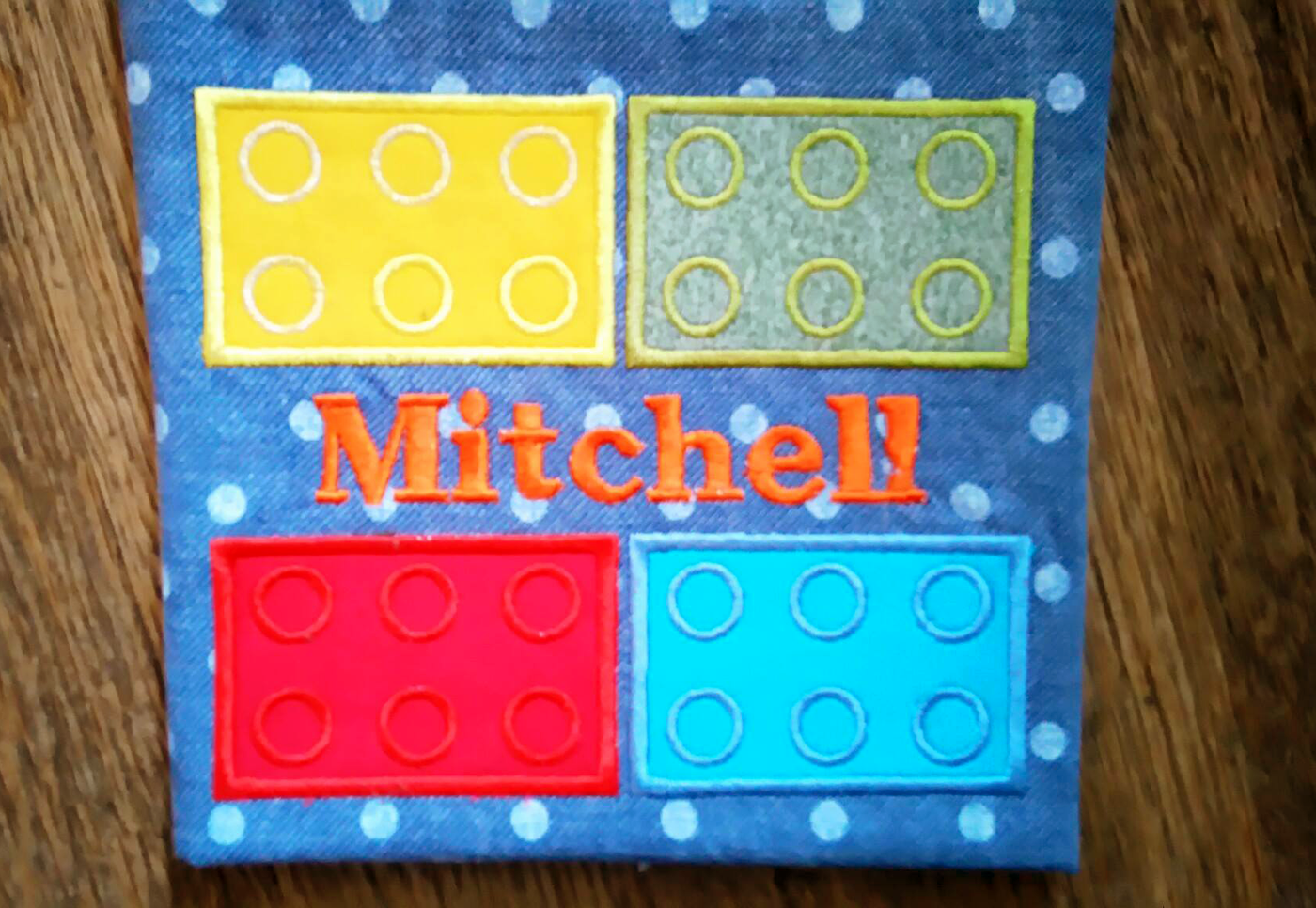 A piece of blue denim fabric with white polkadots. It has an applique design of building bricks on it with a row of bricks at the top and bottom with a split space in between. The customer has added the name "Mitchell" in the split space. (Text is not included with this design.)